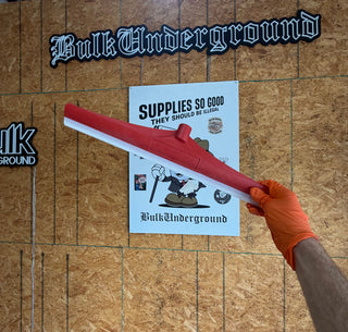 A red and white squeegee held by a hand wearing an orange glove, positioned over a white sign that reads 'Supplies So Good' on a brown oriented-wood panel background.